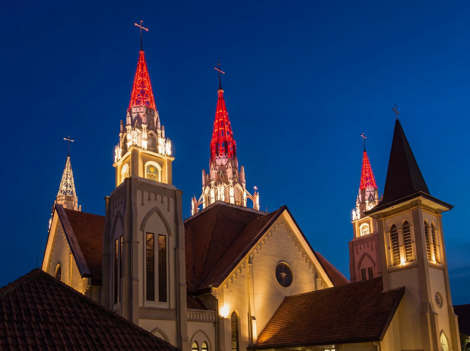 Makassar Cathedral at dusk with red-lit lattice spire crowns above warm white masonry towers, facade lighting by ALTA Integra