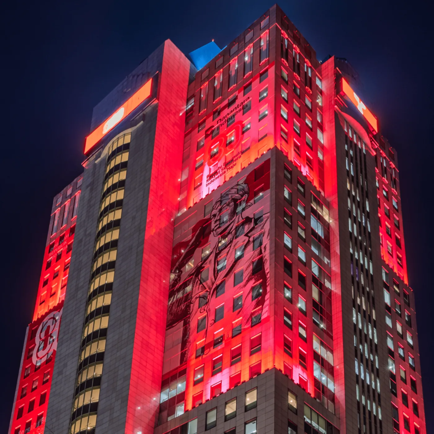 Wisma Danantara facade during the HUT ke-80 treatment, a saturated red field carrying an anniversary lockup, a tagline and a portrait graphic, with warm white on the stair core